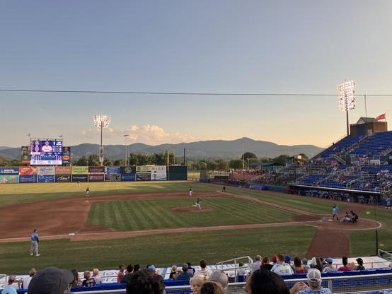 A baseball game is in progress at a stadium, with players on the field and a large scoreboard displaying stats. The background features mountains and a clear sky, with spectators seated in the stands. Stadium lights are illuminated as the sun begins to set.
