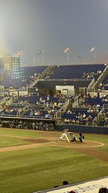 A baseball game in progress at a stadium. The scene shows a batter swinging at a pitch, while the catcher is positioned behind home plate. The stands are filled with spectators, and flags are visible above the seating area. The lighting suggests evening time.