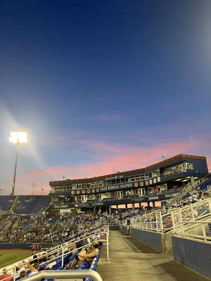 A view of Salem Memorial Ballpark during an event, featuring empty seats and a colorful sunset sky. Stadium lights are on, and a small crowd can be seen in the stands.