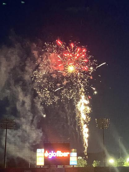 Fireworks explode in the night sky, displaying vibrant colors of red and gold. A scoreboard with the logo "glo fiber" is visible in the foreground, along with stadium lights and a dark backdrop.