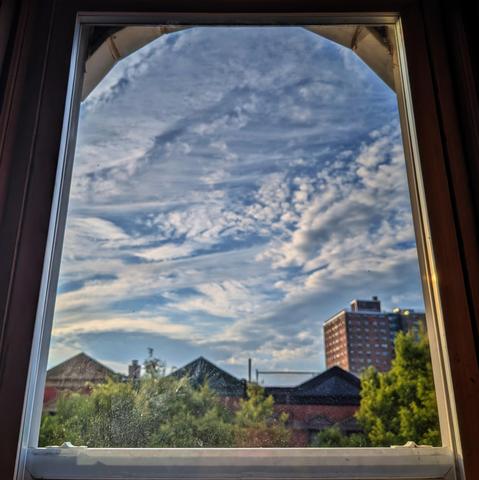 Looking through an arched window ninety minutes after sunrise the blue sky is filled with a swirling tapestry of white clouds. Pointed roofs of Harlem brownstones are silhouetted across the street, and a taller apartment building can be seen in the distance. The green tops of two trees are on the bottom and right.