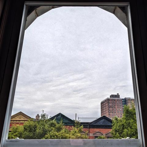 Looking through an arched window three hours after sunrise the sky is filled with a mass of light gray clouds. Pointed roofs of Harlem brownstones with red brickwork are across the street, and a taller apartment building can be seen in the distance. The green tops of two trees are on the bottom and right. 
