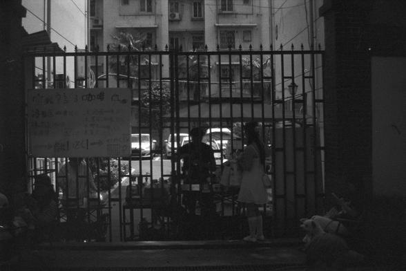 Ilford FP4 Plus 125 (FF)

• English Alt Text: A handwritten sign in Chinese is attached to a gated entrance. Two people are positioned near the gate, one standing and one seated. In the background, parked cars and a building with multiple windows are visible. The sign lists various cakes and baked items repeatedly, such as “coffee,” “oven cake,” and multiple entries of “cake.”
• 中文替代文本：一个手写的中文招牌贴在门口的铁栅栏上，门附近有两个人，一人站着一人坐着。背景中可见停着的车辆和一栋有多个窗户的建筑。招牌上反复列出各类蛋糕和烘焙食品，如“咖啡”、“烤箱蛋糕”和大量“蛋糕”的字样。