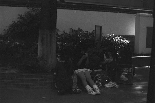 Ilford FP4 Plus 125 (FF)

• English Alt Text: A black-and-white photo of people sitting outdoors on a brick ledge near a tree. The group is partially shaded with sunlight filtering through, and they appear absorbed in their phones or devices. Bags and belongings are placed around them, and the background includes greenery and a plain building wall. The monochrome tone evokes a reflective or timeless atmosphere.
• 中文替代文本：一张黑白照片，几个人坐在室外砖砌的台阶上，旁边有一棵树。阳光从树叶间穿透，给画面带来阴影感，他们似乎都专注于手机或其他设备。周围放有背包和物品，背景是…