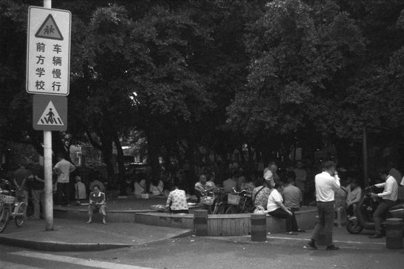 Ilford FP4 Plus 125 (FF)

• English Alt Text: A lively outdoor scene shows people gathered in a shaded, park-like area near a tree-lined street. Some individuals are sitting on benches or ledges, while others stand beside bicycles and motorcycles. A prominent traffic sign on the left warns drivers to slow down near a school zone, with pictograms of children and Chinese text. The environment suggests a casual social gathering in an urban neighborhood, highlighting community life and pedestrian s…
