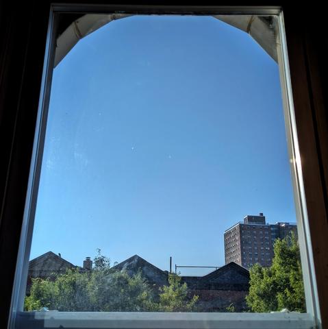Looking through an arched window two hours after sunrise the sky is deep blue and cloudless. Pointed roofs of Harlem brownstones are silhouetted across the street, and a taller apartment building can be seen in the distance. The green tops of two trees are on the bottom and right.