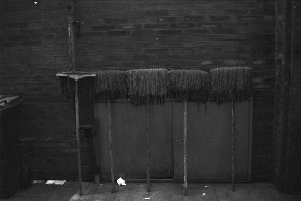 Ilford FP4 Plus 125 (FF)

📝 English Alt Text
Four upright mops are leaning against a red brick wall, their heads resting neatly on a horizontal ledge. To the left, a trash bin sits nearby, and the ground is scattered with bits of debris.

🧼 中文替代文字
四个拖把直立靠在红砖墙上，拖把头整齐地放在横杆上。左侧有一个垃圾桶，地面上散落着一些碎片。