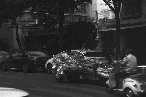 Ilford FP4 Plus 125 (FF)

📝 English Alt Text
A black-and-white street scene featuring a produce cart in the center, filled with fruits and vegetables covered by newspapers and bags. A vendor stands behind the cart, while a scooter rider passes by on the right. Parked cars line the left side, and buildings with balconies rise in the background.

🧺 中文替代文字
这是一张黑白街景照，画面中央是一辆装满蔬果的推车，盖着报纸和塑料袋。一位摊贩站在车后，画面右侧有一名骑电动车的人驶过，左边停着几辆汽车，背景是带阳台的建筑物。