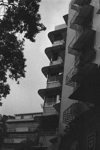 Ilford FP4 Plus 125 (FF)

🏢 English Alt Text
A black-and-white photo of a modern multi-story building featuring staggered balconies with curved roofs and railings. The architectural design emphasizes rhythm and repetition. On the left side, leafy trees soften the scene, creating contrast between the organic and the geometric elements.

🌲 中文替代文字
这是一张黑白照片，展示一栋现代风格的多层建筑，阳台层叠排列，屋顶和栏杆呈弧形，设计富有节奏感。左侧有枝叶茂盛的树木，为画面增添自然元素，与建筑的几何线条形成对比。