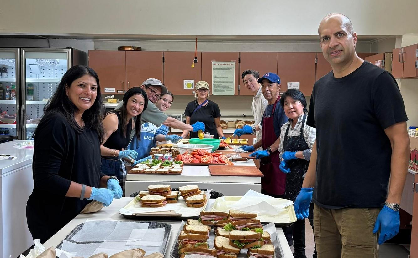 In this photo taken in the Midwest Buddhist Temple kitchen, nine people of various ages, ethniticies, and genders turn to smile for the camera while assembling trays of sandwiches. Picture is from the temple's Facebook page.