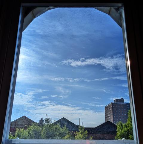Looking through an arched window two hours after sunrise the blue sky is filled with thin, watery white clouds. Pointed roofs of Harlem brownstones are silhouetted across the street, and a taller apartment building can be seen in the distance. The green tops of two trees are on the bottom and right. 
