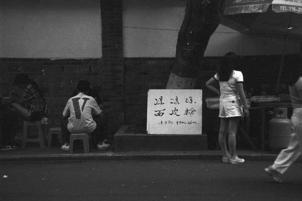 Ilford FP4 Plus 125 (FF)

English alt text:
Outdoor street scene with a group of people gathered around a sidewalk near a tree. In the center, a sign with Chinese characters. The sign promotes different types of cold jelly noodles, suggesting a food stall nearby. The mood appears casual, with individuals sitting and standing around, possibly waiting or socializing.

中文替代文字：
街边场景，一群人围绕在人行道边的树下。中间的牌子暗示附近有凉粉摊位。人们坐着或站着，气氛轻松，可能在等待或聊天。