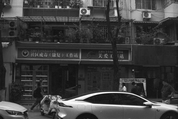 Ilford FP4 Plus 125 (FF)

English alt text:
Street view featuring a white car parked outside a building with two Chinese signs. One sign reads “Community Drug Rehabilitation (Recovery) Workstation” and the other reads “Care Workstation.” A few pedestrians walk nearby, a scooter is parked alongside, and the building has potted plants on the balcony and air conditioning units installed.

中文替代文字：
街景中，一辆白色汽车停在一栋建筑物前，建筑物上挂有两个中文标志，一个写着“社区戒毒（康复）工作站”，另一个写着“关爱工作站”。人行道上有行人经过，旁边停有一辆电动车，建筑阳台上摆放着植物并安装有空调设备。