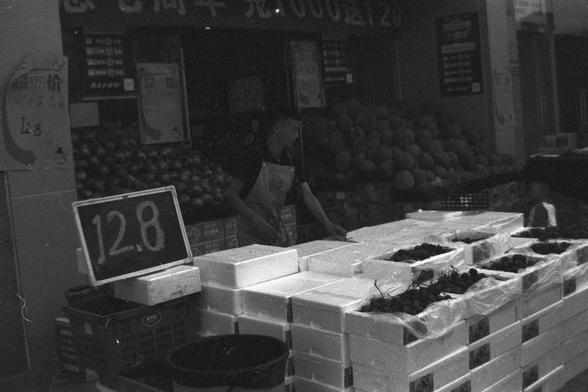 Ilford FP4 Plus 125 (FF)

English alt text:
Black-and-white photo of a bustling market stall. A person stands behind a counter stacked with open white boxes, likely containing produce. A bold handwritten sign marked “12.8” in the foreground suggests a price for the goods. The scene is filled with fruits, vegetables, and various signage, creating a sense of everyday commerce. The grayscale tone gives it a timeless, nostalgic feel.

中文替代文字：
黑白市场景象，一人站在摆满白色箱子的摊位后，箱子里可能装着各类农产品。前方有一个醒目的手写标价牌“12.8”，暗…