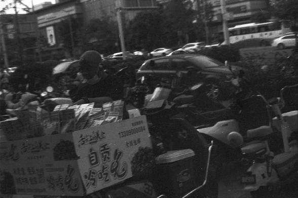 Ilford FP4 Plus 125 (FF)

English alt text:
Black-and-white street scene showing a person standing behind a cart labeled with Chinese characters. The cart displays the phrase “自贡冷吃兔,” suggesting it sells cold spicy rabbit, a specialty from Zigong. Multiple motorcycles are parked nearby. Urban elements like buildings, cars, and a bus in the background convey a busy city atmosphere.

中文替代文字：
黑白街景中，一人站在写有中文标志的小推车后。推车上显示“自贡冷吃兔”，表明售卖的是自贡特色的冷吃兔。周围停放着多辆摩托车，背景中的楼房、汽车和公交车营造出繁忙的都市氛围。