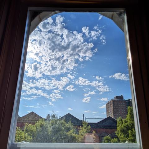 Looking through an arched window two hours after sunrise the blue sky is filled with scattered white clouds. Pointed roofs of Harlem brownstones are silhouetted across the street, and a taller apartment building can be seen in the distance. The green tops of two trees are on the bottom and right.