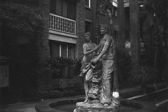 Ilford FP4 Plus 125 (FF)

🗿 English Alt Text
Black and white photo of a statue featuring two figures—one gently placing a hand on the other’s shoulder, while the second figure holds a jar. The statue stands on a pedestal surrounded by a circular fountain in an outdoor courtyard. Behind them is a stone building with decorative lattice windows, adding an old-world charm to the tranquil scene.

🏛️ 中文替代文字
这是一张黑白照片，画面中的雕像由两个人物组成，一人手握水壶，另一人轻触其肩膀。雕像矗立在喷泉中央的基座上，四周环绕着一个圆形水池。背景是一栋石砌建筑，窗户带有装饰性格栅图案，为宁静的庭院增…