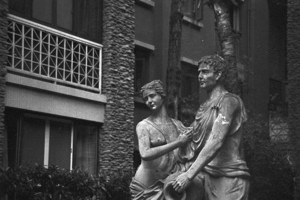 Ilford FP4 Plus 125 (FF)

🧍‍♂️🧍‍♀️ English Alt Text
A black and white image showing two Greco-Roman style statues: a male figure with defined muscles and draped robes gazes ahead, while a female figure beside him appears to look downward, her garment flowing elegantly. They stand in an open courtyard against a backdrop of a textured stone building with balconies featuring geometric metal railings. The atmosphere feels timeless, reminiscent of classical art in a historical European garden.

🎞️ 中…