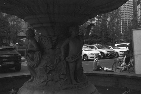 Ilford FP4 Plus 125 (FF)

📝 English Alt Text
A black and white photo of a fountain with two cherub statues at its base, set in an urban plaza. Nearby, one person sits on the fountain’s edge, appearing to read or write, while another stands next to a bicycle. Parked cars and a large blank signboard complete the cityscape in the background.

🧾 中文替代文字
这是一张黑白照片，画面中是一座喷泉，底座上有两个小天使雕像。喷泉位于一个城市广场，一人坐在喷泉边缘，似乎正在阅读或写作，另一人站在自行车旁。背景中有停放的汽车和一个巨大的空白广告牌，构成城市景观。