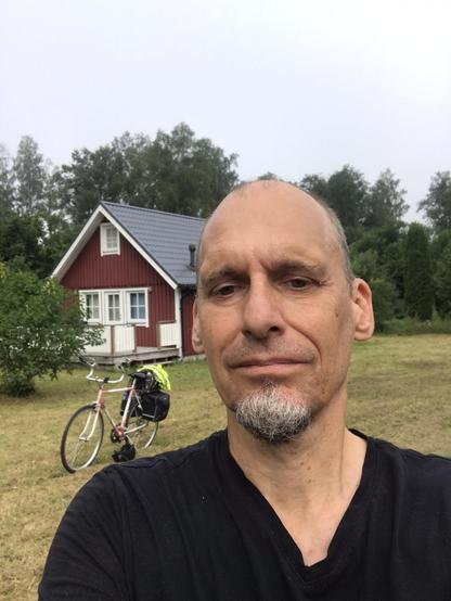 Selfie av Jon Brunberg stående framför ett fritidshus med en packad cykel emellan.