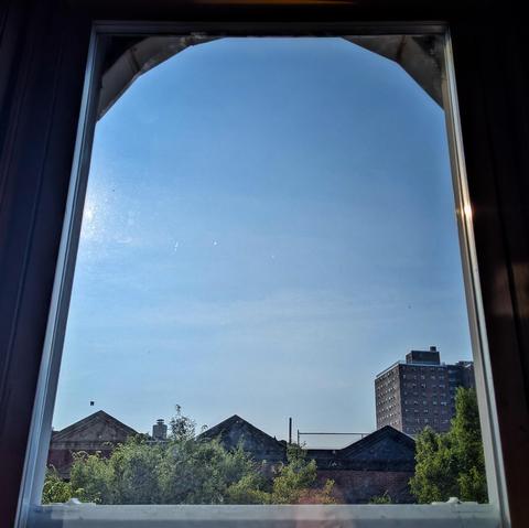 Looking through an arched window two hours after sunrise the blue sky is marked with subtle white clouds, and the Sun is about to enter the frame on the left hand side. Pointed roofs of Harlem brownstones are silhouetted across the street, and a taller apartment building can be seen in the distance. The green tops of two trees are on the bottom and right.