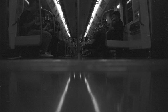 Ilford FP4 Plus 125 (FF)

📝 English Alt Text
A black and white photograph of the inside of a subway or train car, taken from a low angle near the floor. Passengers are seated along both sides—some engaged with their phones, others quietly gazing ahead. Overhead lights cast soft reflections across the glossy floor, adding depth and a cinematic feel to the narrow space.

🖋️ 中文替代文字
一张黑白照片，从靠近地面的低角度拍摄列车车厢内部。乘客分坐在两侧，有人低头看手机，有人沉思望前方。头顶的灯光在光滑的地板上投射出柔和的倒影，使狭长的空间显得格外有层次感与电影感。