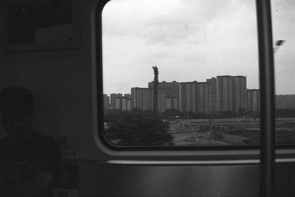 Ilford FP4 Plus 125 (FF)

📝 English Alt Text
A black and white photo taken from inside a train looking out through a window. Outside, a dense urban landscape is visible, featuring multiple tall apartment buildings clustered together. A construction site with machinery and stacked materials occupies the foreground. A passenger is partially visible in the bottom left, adding a candid, contemplative feel to the scene.

🖋️ 中文替代文字
一张从火车车厢内拍摄的黑白照片，透过窗户可见外面的城市景观。多栋高层公寓楼密集排列，前方是一片施工工地，有机械设备和堆放的建材。画面左下角…