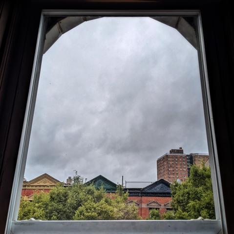 Looking through an arched window three hours after sunrise the sky is filled with gray clouds. Pointed roofs of Harlem brownstones with red brickwork are across the street, and a taller apartment building can be seen in the distance. The green tops of two trees are on the bottom and right. 