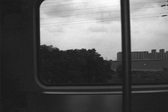 Ilford FP4 Plus 125 (FF)

**English:**
A black and white photograph taken from inside a moving train. The view through the window shows a blurred landscape with trees and buildings in the background, suggesting motion. The window frame is clearly visible, and the scene outside appears to be an urban area with high-rise buildings.

**Chinese:**
一张从行驶中的火车内部拍摄的黑白照片。透过车窗可以看到模糊的景色，背景中有树木和建筑物，暗示着运动感。车窗框清晰可见，窗外的场景看起来是一个有高层建筑的城市地区。