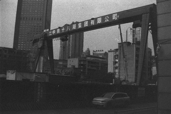 Ilford FP4 Plus 125 (FF)

**English:**
A black and white photograph of an urban construction site. A large gantry crane with Chinese characters on it dominates the scene. The crane is positioned in front of several tall buildings, indicating a cityscape. There is a car on the road in the foreground, and the overall atmosphere suggests ongoing development or construction work.

**Chinese:**
一张城市建筑工地的黑白照片。一台大型的带有中文字的龙门起重机主导了整个场景。起重机位于几座高楼前面，显示出城市天际线。前景的道路上有一辆汽车，整体氛围表明正在进行的开发或建设工作。