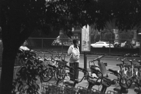 Ilford FP4 Plus 125 (FF)

**English:**
A black and white photograph of a busy urban street scene. The foreground is filled with numerous bicycles parked in rows, suggesting a bike-sharing station or a common parking area for bicycles. A few people are visible, one of whom is standing near a signpost. The background shows a well-lit street with moving vehicles.

**Chinese:**
一张夜晚繁忙的城市街景的黑白照片。前景停满了成排的自行车，暗示着这是一个共享单车站点或自行车的常见停车区。照片中可以看到几个人，其中一人站在一个标志牌附近。背景显示了一条灯火通明的街道，有车辆在移动。