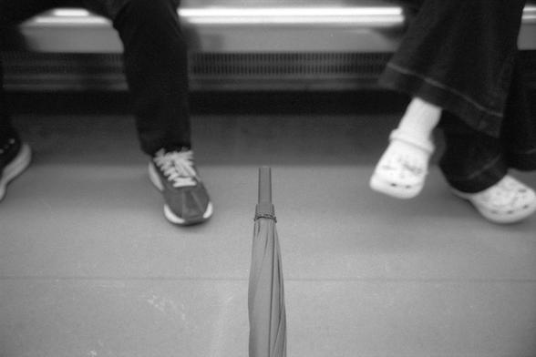 Ilford Pan 400 (FF)

**English:**
A black-and-white photograph taken inside a moving vehicle, possibly a train or bus. The focus is on a closed umbrella standing upright on the floor between the legs of seated passengers.

**Chinese:**
一张在移动车辆内拍摄的黑白照片，可能是在火车或公共汽车上。焦点是一把直立在地板上的收起的雨伞，位于乘客的腿之间。