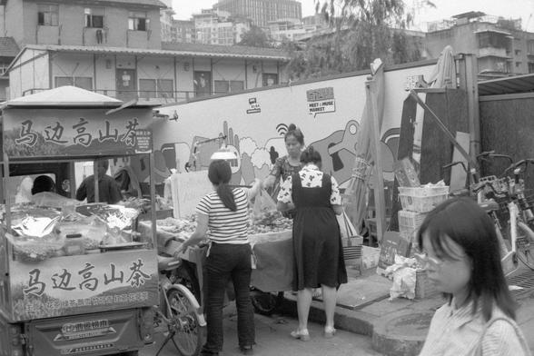 Ilford Pan 400 (FF)

**English:**
A black-and-white photograph showing a street market scene. There are several stalls with various goods on display. Two women are interacting at one of the stalls, which has a sign with Chinese characters. The market appears to be bustling with activity, and there are bicycles and other market items visible in the background.

**Chinese:**
一张黑白照片，展示了一个街头市场的场景。有几个摊位展示着各种商品。两名女子在其中一个摊位互动，该摊位有一个写有中文字的招牌。市场看起来热闹非凡，背景中可以看到自行车和其他市场物品。