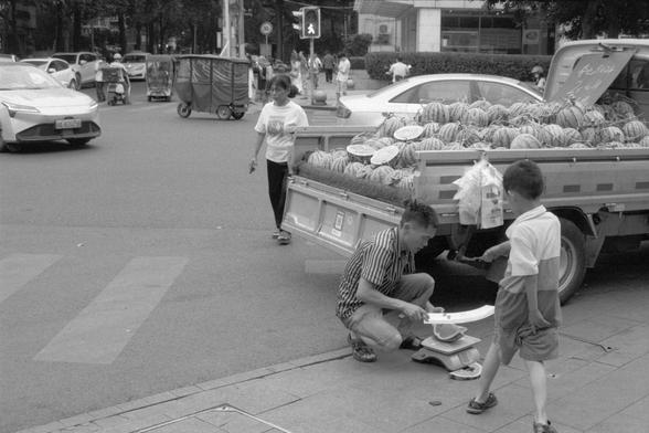 Ilford Pan 400 (FF)

**English:**
A black-and-white photograph depicting a street scene where a man is squatting on the sidewalk next to a weighing scale. A truck loaded with watermelons is parked nearby, and a young boy is standing close to the man. The background shows a busy street with several pedestrians and vehicles, including cars and motorbikes.

**Chinese:**
一张黑白照片，描绘了一个街道场景，一名男子蹲在人行道上，旁边有一个称重秤。附近停着一辆装满西瓜的卡车，一个小男孩站在男子旁边。背景显示了一条繁忙的街道，有几个行人和车辆，包括汽车和摩托车。