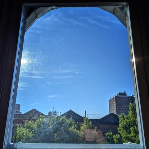 Looking through an arched window two hours after sunrise the deep blue sky is marked with subtle horizontal slivers of white cloud. Pointed roofs of Harlem brownstones are silhouetted across the street, and a taller apartment building can be seen in the distance. The green tops of two trees are on the bottom and right. 