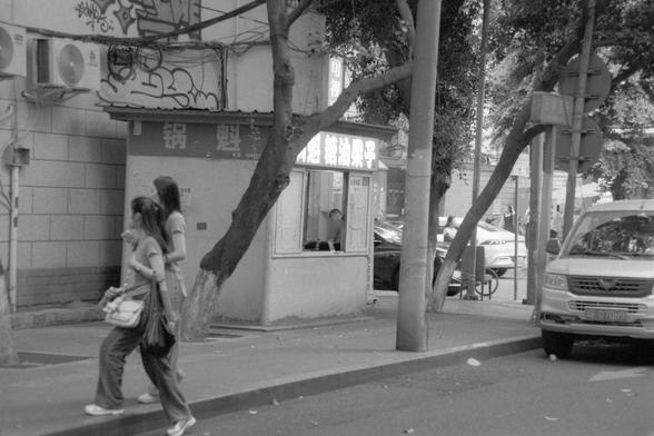 Ilford Pan 400 (FF)

---

📝 English Alt Text:
A black and white street photograph showing two pedestrians walking past a small building with a service window. Above the window is a sign with Chinese characters that read “锅魁糖油果子,” referring to a type of fried sweet snack. A van with the license plate “F Q70H9” is parked on the right side of the street. Trees and a utility pole are visible in the foreground, adding texture to the urban scene.

🌏 中文替代文字：
一张黑白街景照，画面中两名行人经过一个带服务窗口的小建筑。窗口上方悬挂着写有“锅魁糖油…