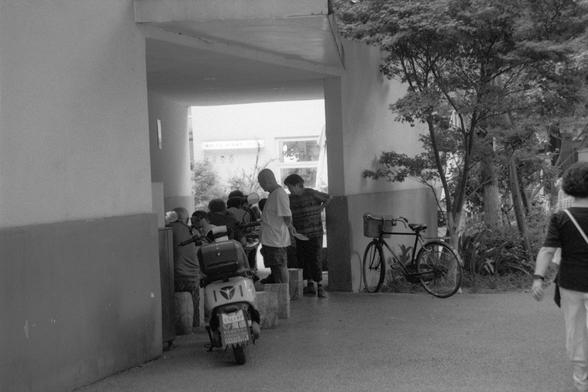 Ilford Pan 400 (FF)

---

📝 English Alt Text:
A black and white photo depicting a group of people gathered under a covered walkway beside a building. A scooter with a visible license plate is parked in the foreground, and a bicycle leans against the wall on the right. On the far side, a storefront sign reads “WHITE NIGHT.” Trees and plants line the right side of the scene, softening the urban atmosphere. The image evokes a moment of casual social interaction in a quiet residential area.

🌏 中文替代…