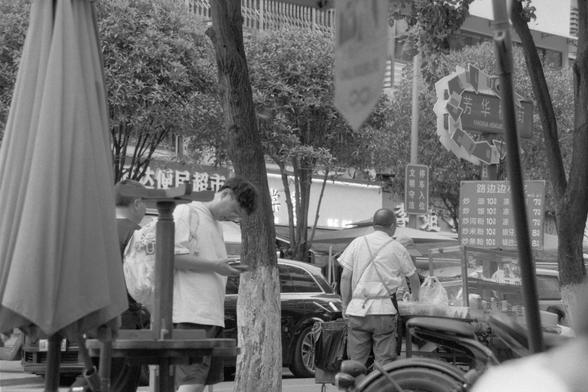 Ilford Pan 400 (FF)
---

📝 English Alt Text:
A black and white photograph of an urban street scene featuring several people. One person stands and looks down at a mobile phone, while another walks away in the distance. Behind them are buildings, trees, and signage including one that reads “FANGHUA HENGJIE.” A poster listing food items and prices is visible near a storefront. The atmosphere feels casual, capturing a slice of daily life in a city environment.

🌏 中文替代文字：
一张黑白城市街景照，画面中几位人物包括一位低头看手机…