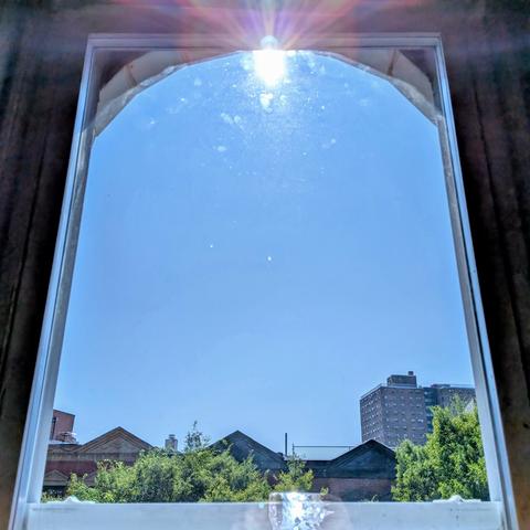 Looking through an arched window four hours after sunrise the sky is bright blue and cloudless. The sun is directly above, creating a lens flare at the top of the frame. Pointed roofs of Harlem brownstones are silhouetted across the street, and a taller apartment building can be seen in the distance. The green tops of two trees are on the bottom and right. 