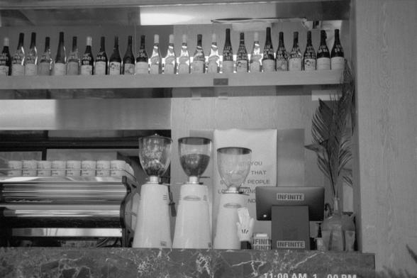 Ilford Pan 400 (FF)

English alt text:
Black and white photo of a café or bar counter. A variety of wine bottles are arranged on the top shelf. Beneath, two coffee grinders and a coffee machine sit on the marble countertop. A partially visible sign hangs behind the grinders. Two small signs with the word “INFINITE” are placed on the counter. In the bottom right corner, the time range “11:00 AM - 1:00 PM” is displayed.

中文替代文字：
这是一张黑白照片，展示一个咖啡馆或酒吧的柜台。顶层架子上整齐地排列着各种葡萄酒瓶。下方的大理石台面上放着两个咖啡研磨机和一台咖啡机。研磨…
