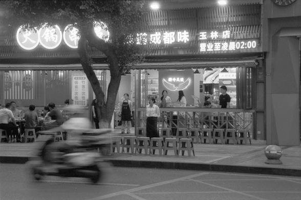 Ilford Pan 400 (FF)

English alt text:
Nighttime street scene in front of a brightly lit Chinese restaurant. Neon signs and a large storefront banner in Chinese indicate the restaurant’s name and that it’s open until 2:00 AM. Several patrons are seated at outdoor tables enjoying food, while others stand and talk near the entrance. A motorbike passes in the foreground with visible motion blur. The lively atmosphere contrasts with the dark surroundings.

中文替代文字：
夜晚街头，一家灯光明亮的中式餐厅门前。霓虹灯和中文横幅展示餐厅名称，…