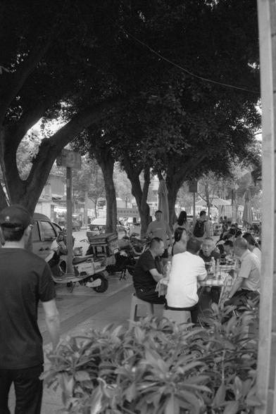Ilford Pan 400 (FF)

English alt text:
Outdoor dining scene under tree shade, with people sitting at checkered-tablecloth tables, enjoying food and conversation. Scooters are parked nearby, enhancing the casual urban vibe. The atmosphere feels communal and relaxed, suggesting a friendly social gathering, possibly in a public square or patio.

中文替代文字：
树荫下的户外用餐场景，人们围坐在铺有格子桌布的餐桌旁，享受美食与交流。周围停放着几辆踏板车，营造出轻松的城市氛围。整体感觉亲切自在，似乎是在广场或露台上的友好聚会。