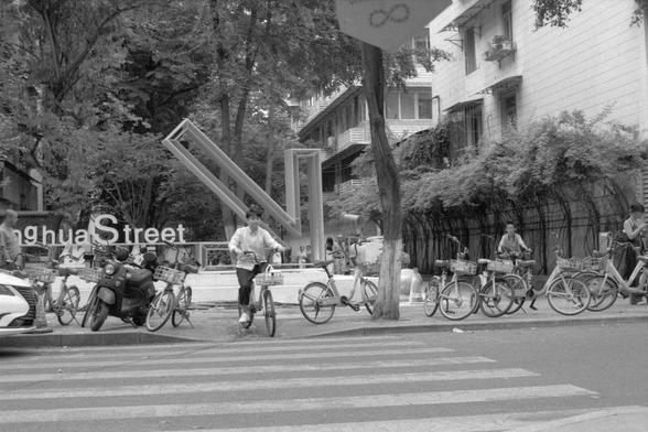 Ilford Pan 400 (FF)

English alt text: Black and white street scene featuring several bicycles parked along a sidewalk. A cyclist rides across a pedestrian crosswalk. In the background, a large sign with a modern sculpture nearby. Trees and buildings frame the urban setting.

中文替代文字：黑白街景照片，几辆自行车停在人行道边。一名骑自行车的人正穿过人行横道。背景中可见大型标志和一个现代雕塑。周围是树木和建筑，呈现出都市氛围。