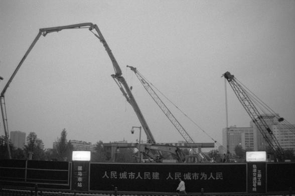 Ilford Pan 400 (FF)

English alt text: A construction site featuring towering cranes and heavy machinery. In the foreground, a fence displays bold Chinese characters that read: “The people’s city is built by the people, the people’s city is for the people.” Behind the fence, buildings and trees line the horizon beneath a cloudy sky.

中文替代文字：一个大型施工现场，几台高大的起重机和重型机械正在运作。画面前方的围栏上写着醒目的标语：“人民城市人民建，人民城市为人民”。远处是树木和建筑，天空布满云层。