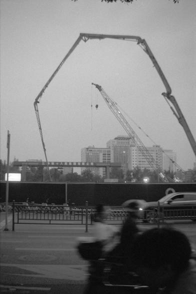Ilford Pan 400 (FF)

English alt text: Black and white photo of an urban construction site with two tall cranes and a concrete pump forming a triangular shape in the sky. In the foreground, blurred motion suggests cyclists or pedestrians crossing the street, with a car passing nearby. Buildings and signage appear in the background, hinting at a bustling city environment.

中文替代文字：这是一张黑白城市施工现场照片，两台高大的起重机与一台混凝土泵在空中形成三角形结构。前景中有行人或骑自行车者的模糊动态，以及一辆驶过的汽车。背景为建筑与标识，呈现出繁忙的都市氛围。