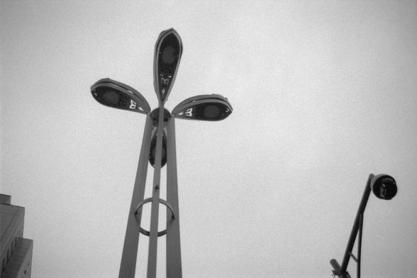 Ilford Pan 400 (FF)

English alt text: Black and white photo featuring a tall streetlight with four angular lamp heads forming a cross-like structure at the top. Another similar streetlight stands to the right, while a building is partially visible on the left side. The sky above is cloudy, lending a moody and industrial atmosphere to the scene.

中文替代文字：黑白照片中，一盏高大的路灯顶端有四个灯头，呈十字形排列。右侧还有一盏类似的路灯，左边隐约可见一栋建筑。天空多云，为画面增添了阴郁的工业气息。