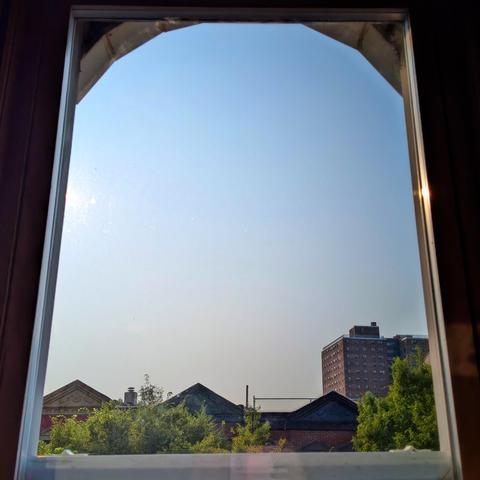 Looking through an arched window two hours after sunrise the cloudless sky fades from light gray at the horizon to palest blue up above. Pointed roofs of Harlem brownstones are silhouetted across the street, and a taller apartment building can be seen in the distance. The green tops of two trees are on the bottom and right.