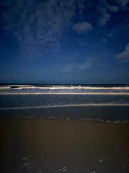 Nighttime beach scene with dark sand, gentle waves lapping at the shore, and a starry sky partially covered by clouds.
