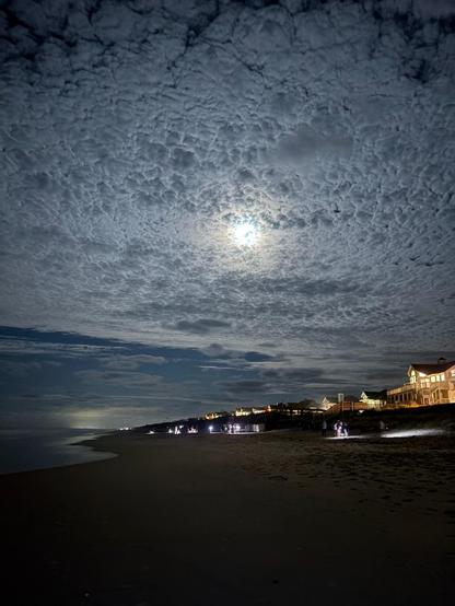 A moonlit beach scene at night, featuring a textured cloud-filled sky above. The coastline is lined with illuminated buildings, and a few people can be seen along the shoreline, some using flashlights. The ocean reflects the moonlight, creating a serene view. 