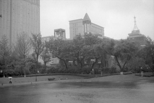 Ilford Pan 400 (FF)

**English:**
A black and white photograph of an urban park scene. The foreground features a large open grassy area with a few trees scattered around. In the background, there are several large buildings, including one with a clock tower and another with a spire. The park appears to be a peaceful retreat within a bustling city.

**Chinese:**
一张黑白照片，展示了一个城市公园的场景。前景是一片宽阔的草地，周围散落着几棵树。背景中有几座大型建筑，其中一座带有钟楼，另一座带有尖顶。这个公园看起来是繁华城市中的一片宁静绿洲。